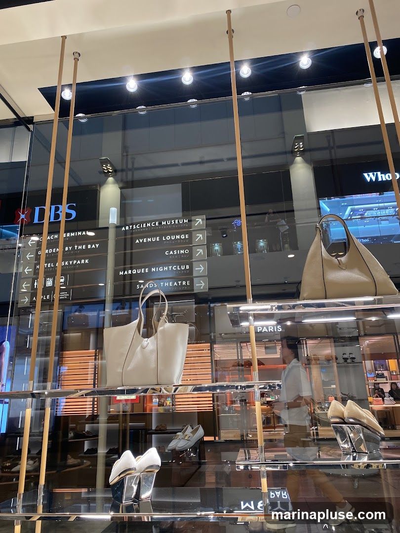 A store window display featuring handbags and shoes. In the background, directional signs indicate various attractions such as ArtScience Museum, Avenue Lounge, Casino, Marquee Nightclub, and Sands Theatre. The store name 'PARIS' is visible.
