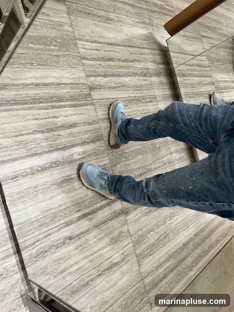 A person is sitting or standing on a floor with a marble-like pattern. They are wearing light blue sneakers with tan soles and distressed denim jeans with paint splatters. The image is taken from a high angle, focusing on the lower body. Mirrors are visible in the background, reflecting the scene.