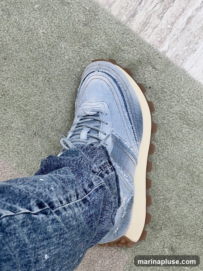 A close-up shot of a person wearing distressed denim jeans and denim sneakers. The sneakers have a chunky sole and a patchwork denim design. The jeans also appear to be distressed with paint splatters. The background is a textured carpet and a marble-like surface.