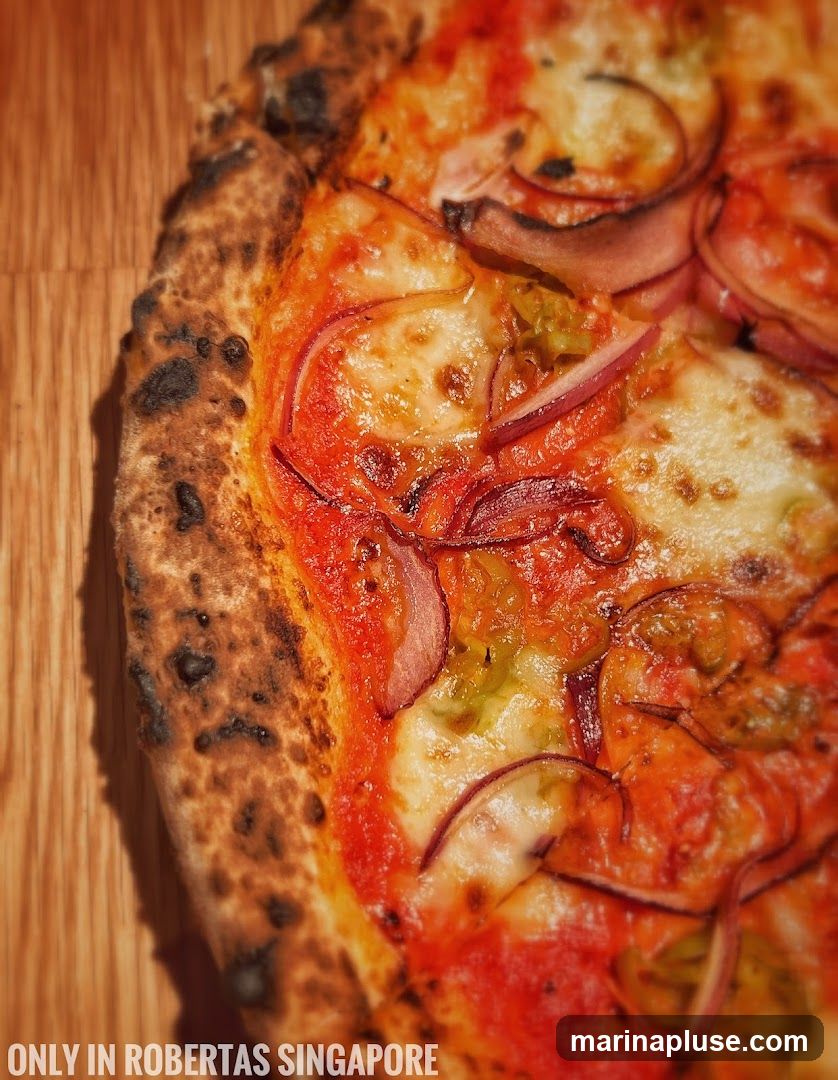 A close-up shot of a pizza with a charred crust, topped with tomato sauce, melted cheese, red onion slices, and green peppers. The pizza is placed on a wooden surface. Text at the bottom reads "ONLY IN ROBERTAS SINGAPORE".