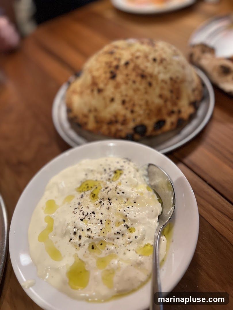 A close-up shot of a white bowl filled with a creamy dip, drizzled with olive oil and sprinkled with black pepper. A spoon rests in the bowl. In the background, a round, golden-brown bread with charred spots sits on a plate. The food is arranged on a wooden table.