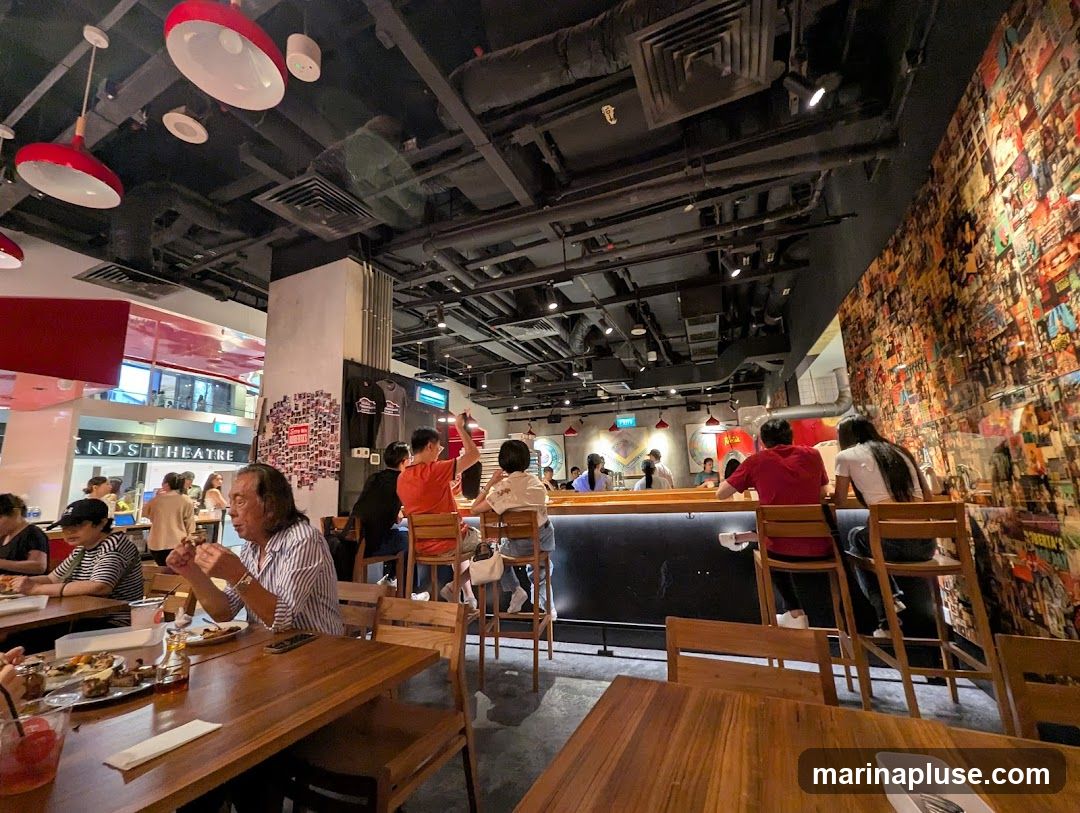 The image shows the interior of a restaurant with several patrons seated at tables and a bar. The walls are decorated with a collage of images and posters. Red pendant lights hang from the black ceiling, which also features exposed ductwork.