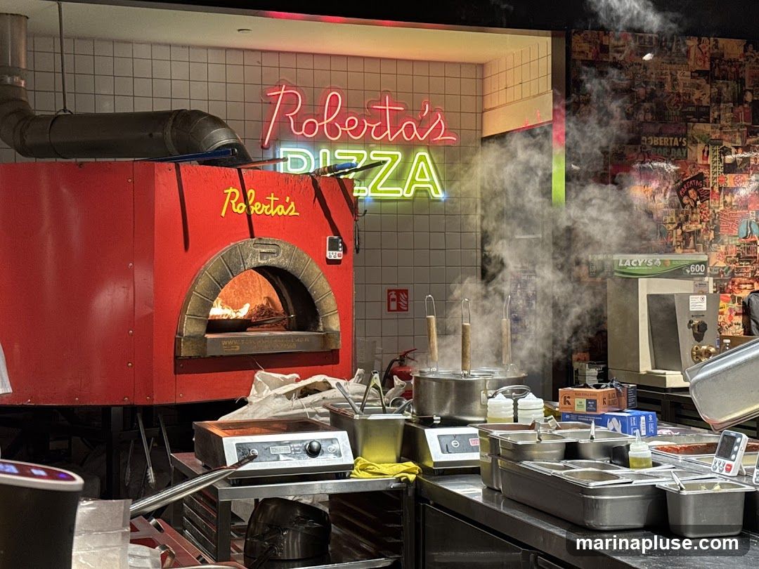 A red pizza oven is visible with a pizza inside. Steam rises from pots on a counter in front of the oven. Neon signs spell out "Roberta's PIZZA". The wall behind the oven is tiled, and the wall to the right is covered in a collage of images and text.
