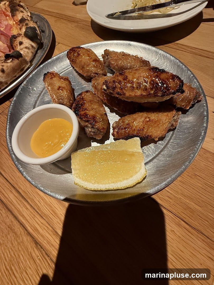 A metal plate holds several cooked chicken wings, a small white dish of yellow dipping sauce, and a slice of lemon. A portion of a pizza is visible on another metal plate to the left. A white plate with food remnants and cutlery is in the background. The food is arranged on a wooden table.