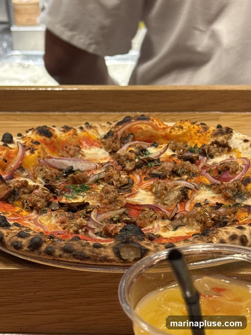A close-up shot of a pizza with sausage, red onions, and herbs, served on a wooden board. In the foreground, a plastic cup filled with a yellow beverage and ice, with a black straw, is visible. The background shows a person in a white uniform, likely a chef or server, in a kitchen setting.