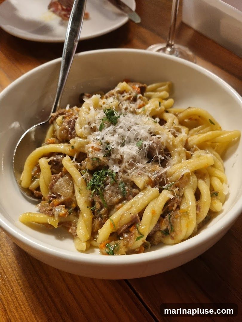 A close-up shot of a bowl of pasta with a meat sauce and grated cheese. A spoon is in the bowl. In the background, out of focus, is a plate with sliced meat and a wine glass.