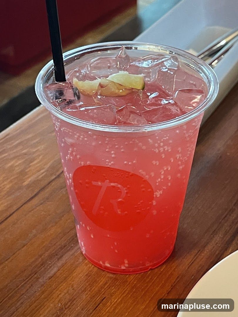 A close-up shot of a pink, bubbly beverage in a clear plastic cup. The drink is filled with ice and garnished with two small wedges of lime. A black straw is inserted into the cup. The cup has a red, circular logo with a white stylized letter 'R' on it. The cup sits on a wooden surface. In the background, a white tray with silverware is visible.