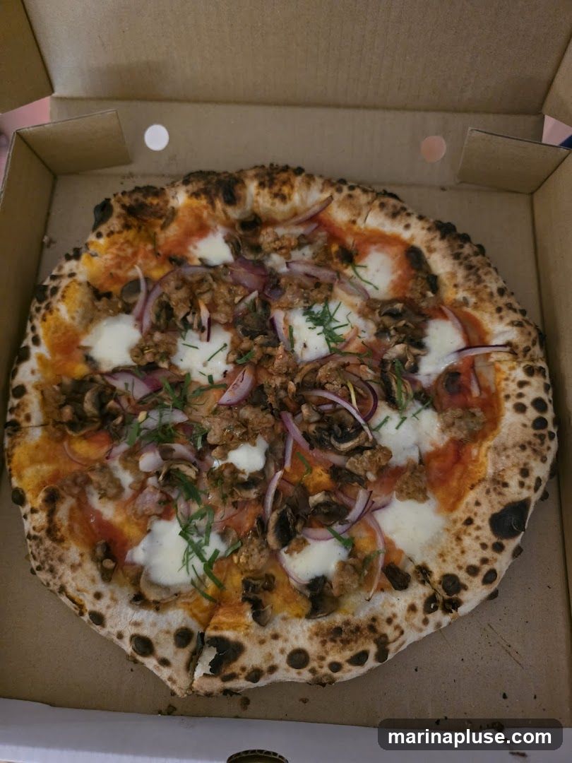 A close-up shot of a pizza in a cardboard box. The pizza has a crust with charring, tomato sauce, mozzarella cheese, sliced mushrooms, red onions, and crumbled meat. It is garnished with fresh herbs.