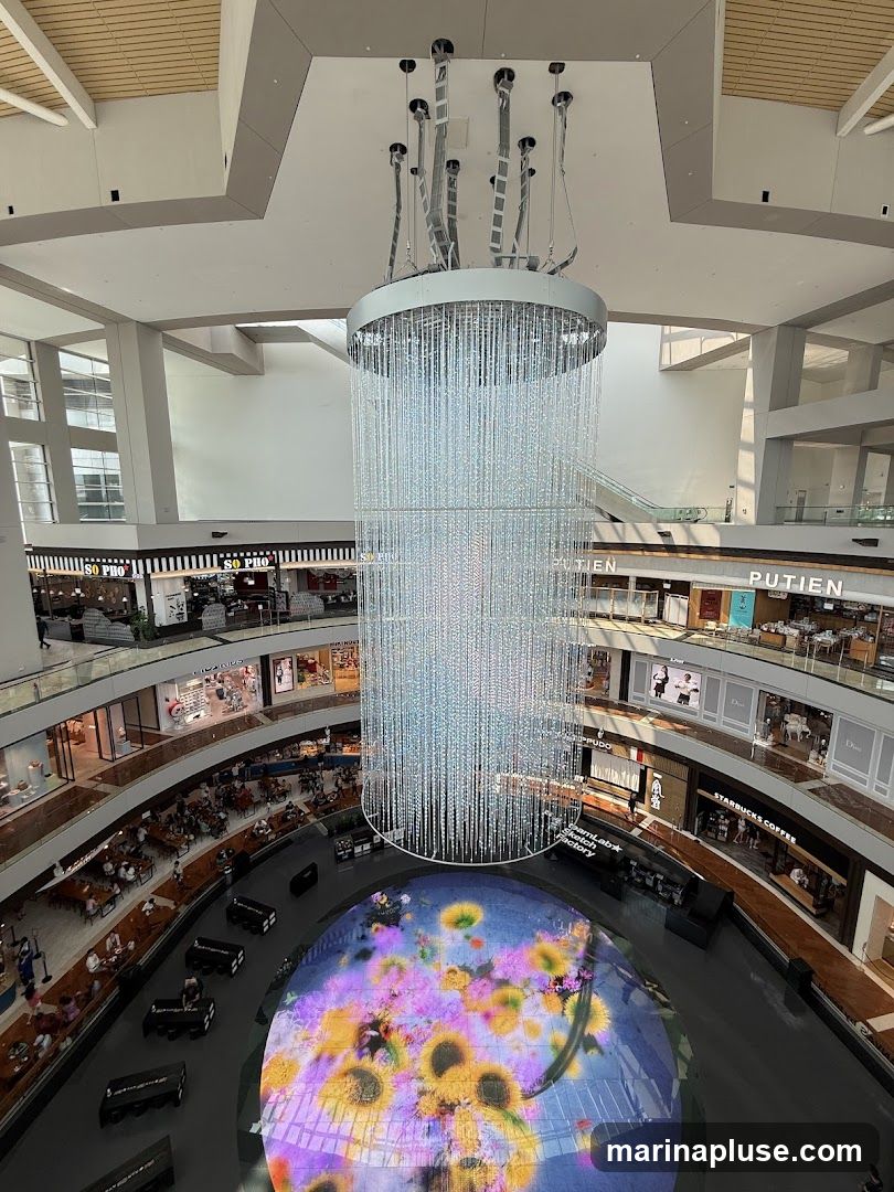 An aerial view of a modern indoor shopping mall atrium. A large, multi-tiered crystal chandelier hangs from the ceiling. Below, a circular digital display on the floor shows a vibrant floral pattern with sunflowers and purple flowers. The mall features multiple levels with shops and restaurants visible, including a Starbucks. People are seated at tables in dining areas and walking around. The architecture is contemporary with clean lines and natural light filtering in from large windows.