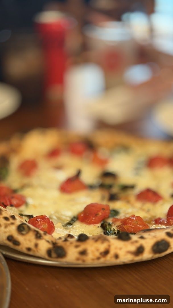A close-up shot of a pizza topped with cherry tomatoes and melted cheese, with a slightly charred crust. In the blurred background, there are red and white cups and people dining.