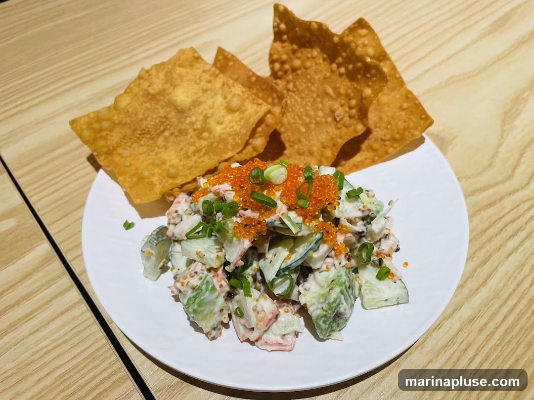 A white plate holds a serving of crab salad, which appears to be made with imitation crab meat, cucumber, and a creamy dressing. It is topped with bright orange tobiko (flying fish roe) and chopped green onions. Two large, crispy wonton chips are placed behind the salad. The dish is presented on a light-colored wooden surface.