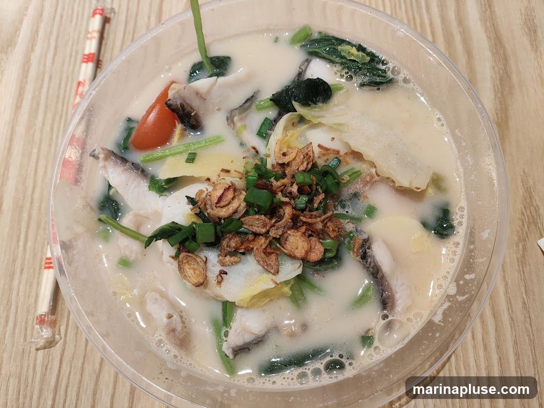 A close-up overhead shot of a bowl of fish soup. The soup is milky white and contains chunks of fish, leafy green vegetables, slices of ginger, a cherry tomato, and is garnished with fried shallots and chopped scallions. A pair of chopsticks in a wrapper is visible to the left of the bowl.