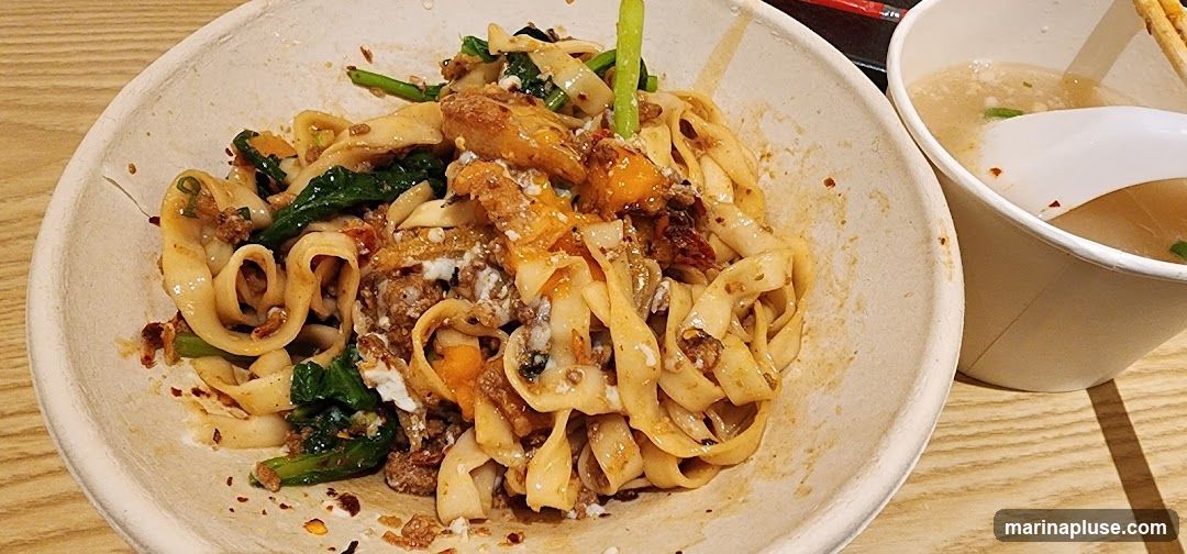 A close-up shot of a bowl of noodles mixed with ground meat, green vegetables, and what appears to be some sort of sauce or topping. A side of broth in a paper cup is visible to the right, with a white spoon inside. The food is served in a light-colored, textured bowl on a wooden surface.