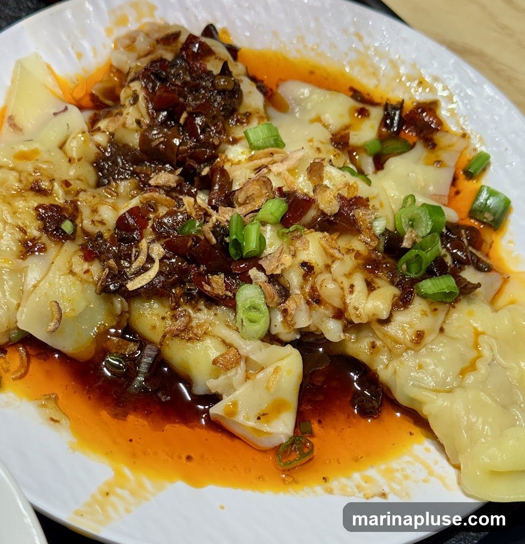 A plate of wontons in a spicy chili oil sauce, garnished with chopped scallions and fried garlic.