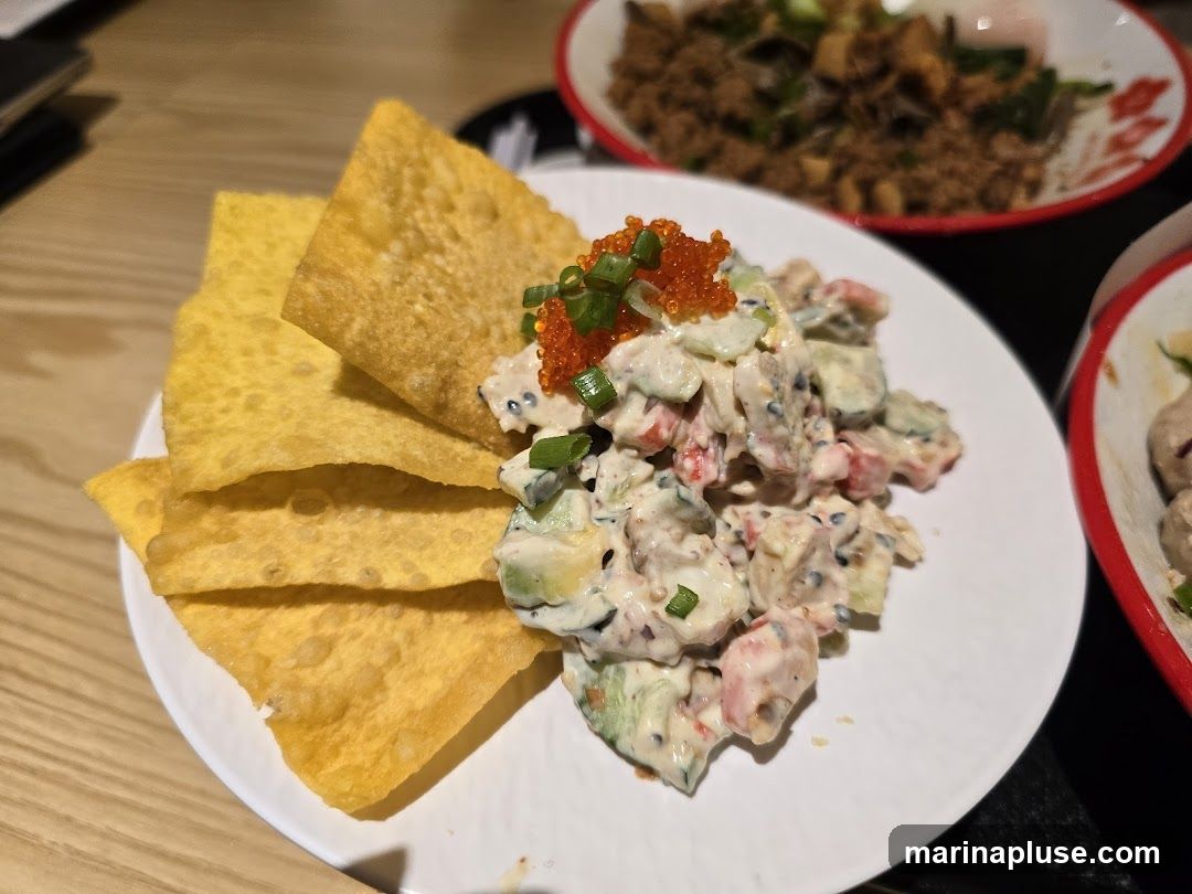 A close-up shot of a creamy avocado salad served with crispy wonton chips. The salad is topped with bright orange roe and chopped green onions. In the background, other dishes are visible, including a bowl of what appears to be minced meat with vegetables and another dish with meatballs. The food is presented on a white plate with a red rim, set on a dark surface, with a wooden table visible in the foreground.