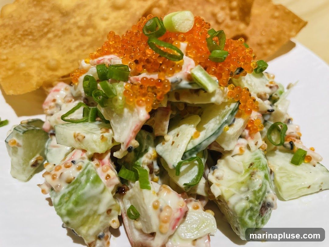 A close-up shot of a creamy crab salad topped with bright orange tobiko (flying fish roe) and chopped green onions. Slices of cucumber and imitation crab meat are visible throughout the salad, which is coated in a creamy dressing. Two crispy wonton chips are placed behind the salad.