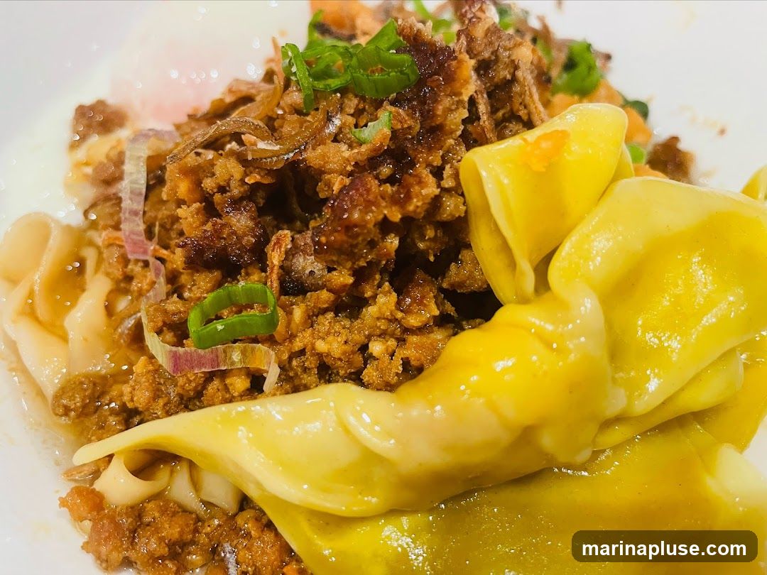 A close-up shot of a dish featuring wide noodles, a generous portion of minced meat, crispy fried shallots, and green onions. There are also some dumplings visible in the foreground. A soft-boiled egg yolk is partially visible in the background.