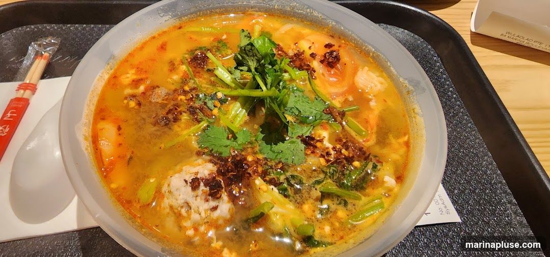 A close-up shot of a bowl of Tom Yum soup, featuring a rich, orange broth filled with visible ingredients such as shrimp, a large meatball, and fresh cilantro. The soup is garnished with chili flakes and green vegetables. A white spoon and a red packet containing chopsticks are placed to the left of the bowl on a black tray. A small white slip of paper with text is visible under the bowl.