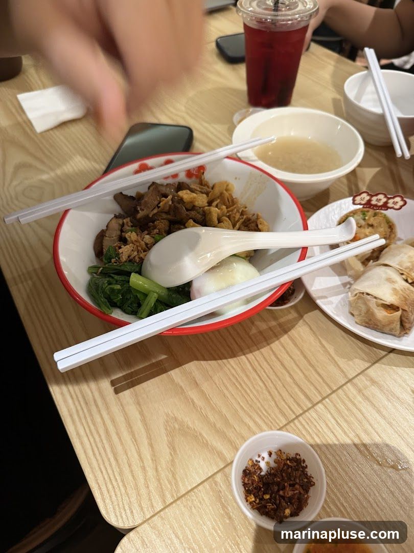 A close-up shot of a bowl of noodles topped with meat and greens, accompanied by a soft-boiled egg. Chopsticks and a spoon rest on the bowl. In the background, a glass of red beverage, a small bowl of soup, and another plate with food items are visible on a wooden table. A small dish of chili flakes is in the foreground.