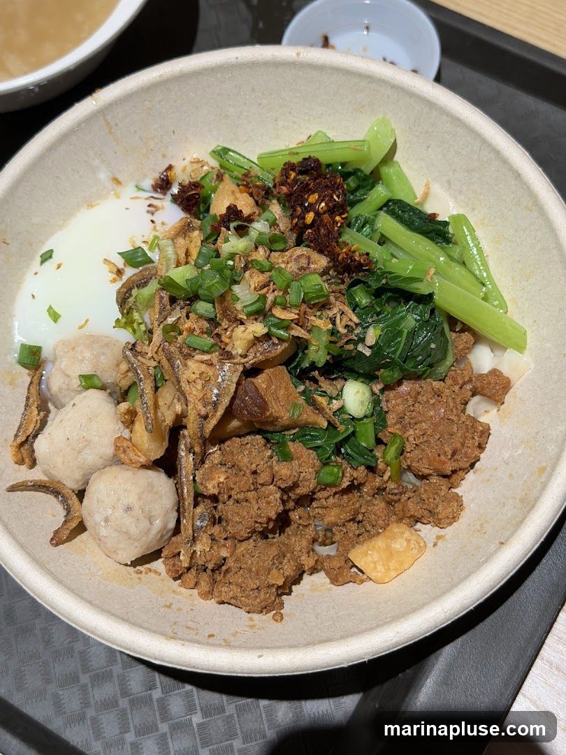 A close-up shot of a bowl of noodles topped with minced meat, meatballs, fried anchovies, green vegetables, a soft-boiled egg, and chili flakes. The bowl is made of a light-colored, textured material and sits on a dark gray tray. A small white bowl with some liquid is visible in the upper right corner.