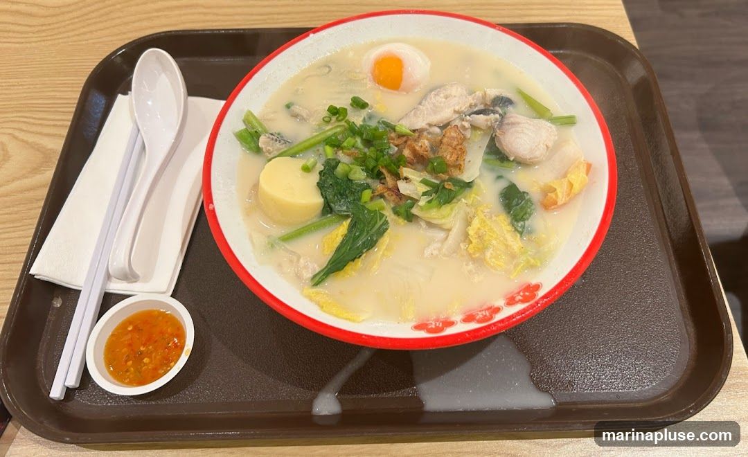 A bowl of fish soup with chunks of fish, soft tofu, leafy green vegetables, and a soft-boiled egg is served on a brown tray. A small dish of chili sauce and a set of chopsticks and a spoon are placed on a white napkin to the side. The soup appears creamy and is garnished with chopped scallions and fried shallots.