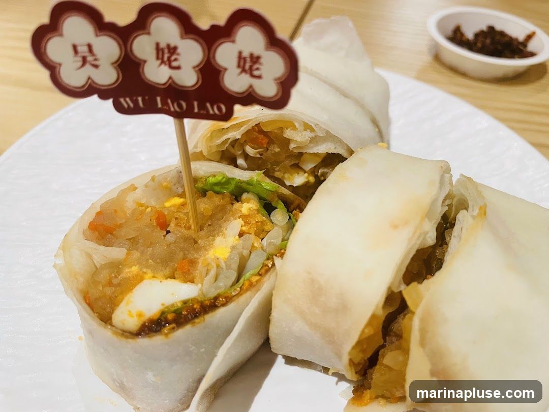 Three spring rolls are presented on a white plate. A skewer with a decorative tag bearing Chinese characters and the text "WU LAO LAO" is inserted into one of the rolls. A small dish of chili paste is visible in the upper right corner.