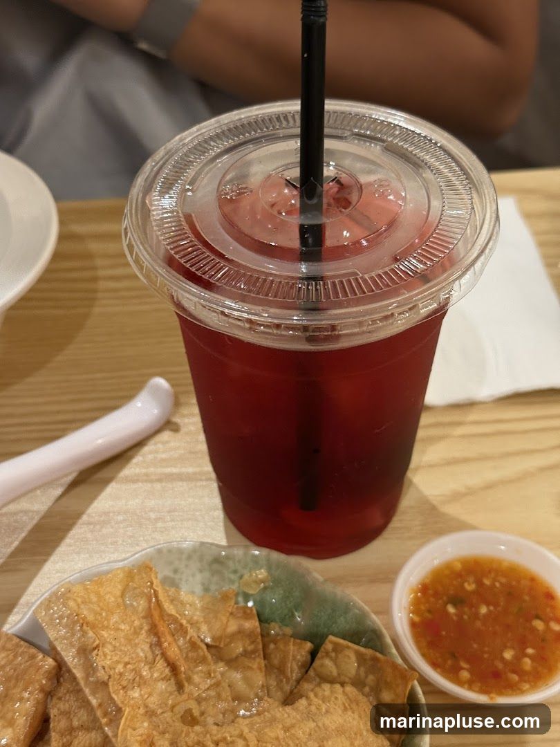 A close-up shot of a red beverage in a clear plastic cup with a black straw, placed on a wooden table. In the foreground, a plate of fried appetizers is visible, along with a small dish of dipping sauce.