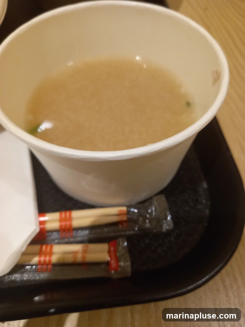 A close-up shot of a bowl of miso soup with chopsticks on a black tray. The soup is a light brown color with some green specks. The chopsticks are individually wrapped in plastic with red bands.