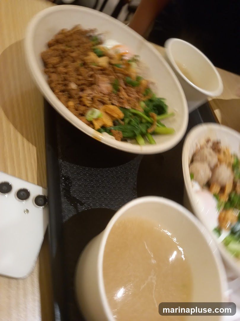 Three bowls are visible on a table. One bowl contains rice with minced meat, greens, and a fried egg. Another bowl has meatballs and greens. The third bowl contains a light-colored soup. A white smartphone is also on the table.