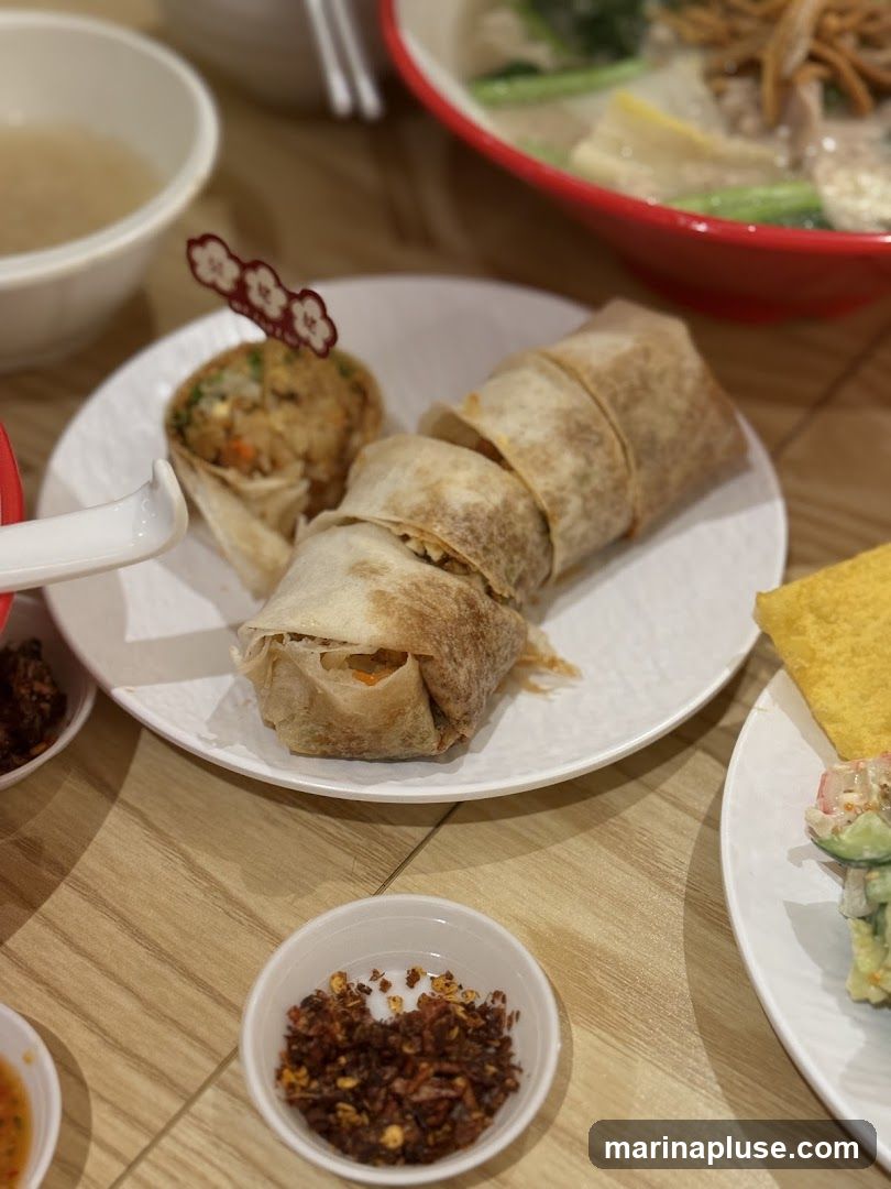 A close-up shot of a plate of sliced spring rolls and a small round item topped with a decorative skewer. In the background, a bowl of soup and another dish with salad are visible. A small dish of chili flakes is in the foreground.