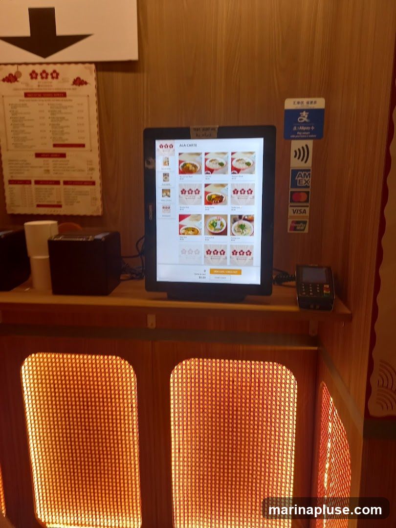 A digital ordering kiosk displays a menu with various food items. To the left, a physical menu board is visible. Payment terminals and a credit card reader are also present. The area is illuminated by warm, patterned lighting.