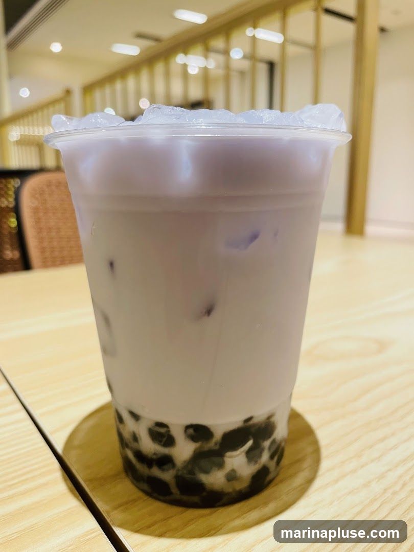 A close-up shot of a taro bubble tea drink in a clear plastic cup, filled with ice and tapioca pearls at the bottom. The drink is a light purple color and is placed on a wooden table. The background shows a blurred interior with wooden accents and lighting fixtures.