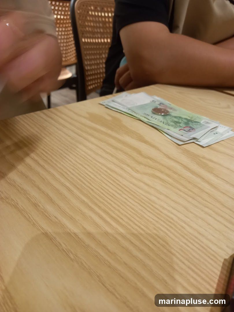 A close-up shot of Singaporean dollar bills and a coin fanned out on a light-colored wooden table. The image is taken from a slightly elevated angle, focusing on the currency. In the background, blurred figures of people and chairs are visible, suggesting a dining or social setting.