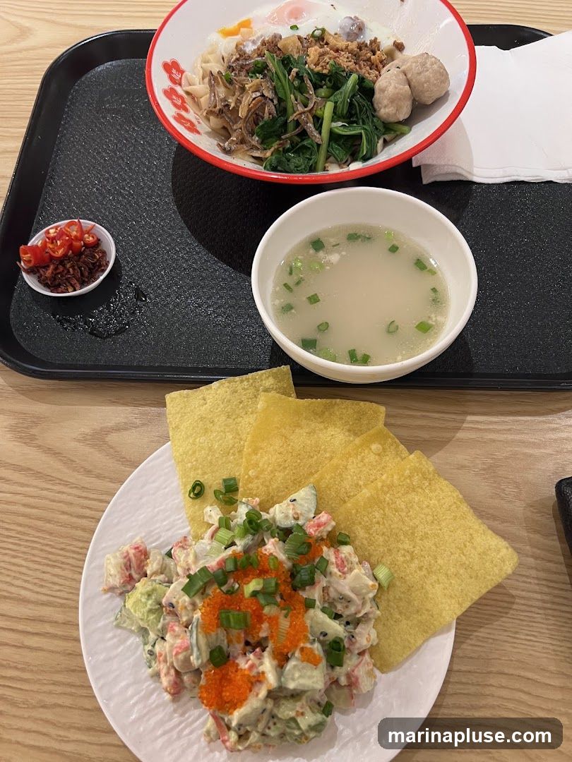 A close-up shot of a meal served on a black tray. The meal includes a bowl of noodles topped with greens, fried shallots, two eggs, and meatballs. To the side is a small dish of chopped chilies and fried shallots. Another bowl contains a clear broth with chopped green onions. In the foreground, a white plate holds a salad with imitation crab, cucumber, and avocado, garnished with green onions and orange roe, accompanied by several fried wonton wrappers.