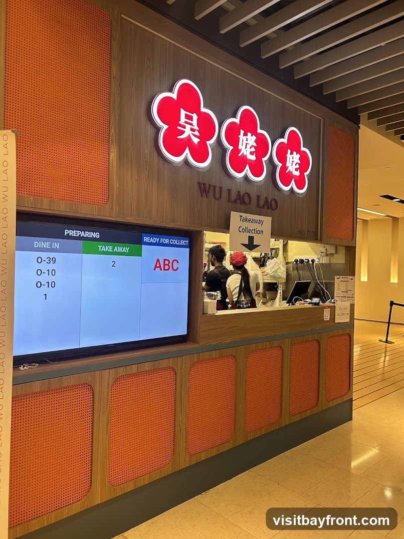 A takeaway counter for Wu Lao Lao, a food establishment. A digital screen displays order statuses for dine-in and takeaway. The counter has a wooden facade with orange textured panels. The brand's logo, featuring red floral shapes with Chinese characters, is prominently displayed above the counter. Staff members are visible behind the counter preparing orders.