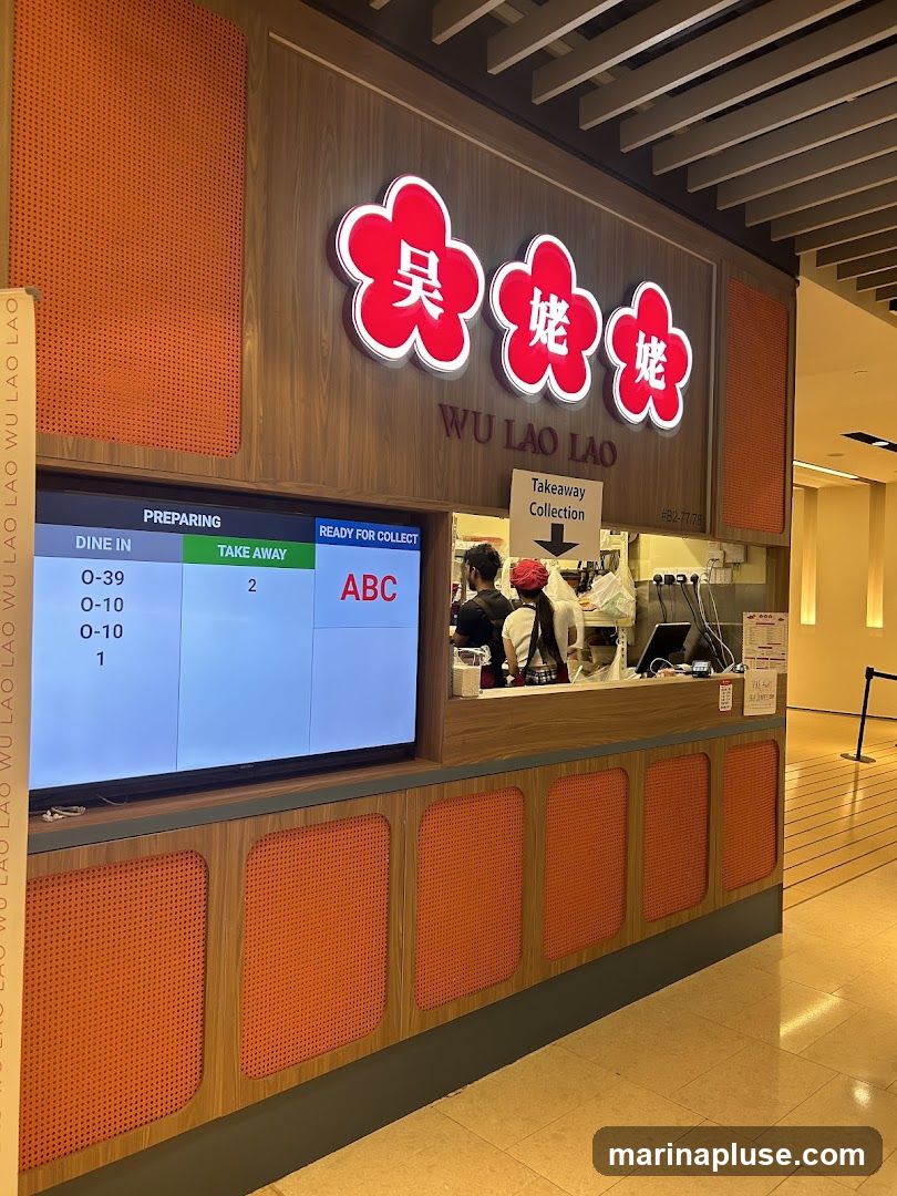 A takeaway counter for Wu Lao Lao, a food establishment. A digital screen displays order statuses for dine-in and takeaway. The counter has a wooden facade with orange textured panels. The brand's logo, featuring red floral shapes with Chinese characters, is prominently displayed above the counter. Staff members are visible behind the counter preparing orders.