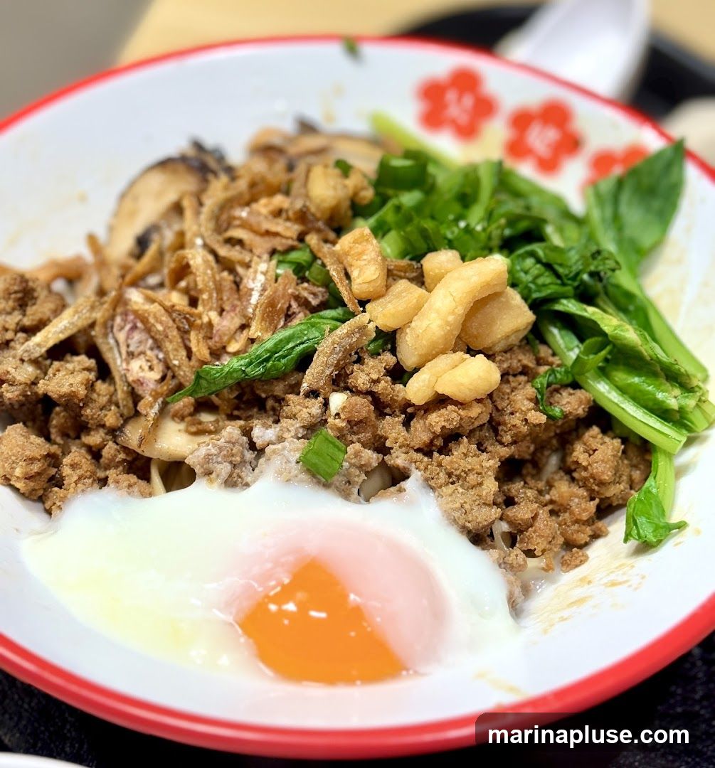 A close-up shot of a bowl of noodles topped with minced meat, fried anchovies, mushrooms, greens, crispy pork rinds, and a soft-boiled egg with a runny yolk. The bowl has a red rim with a floral pattern.
