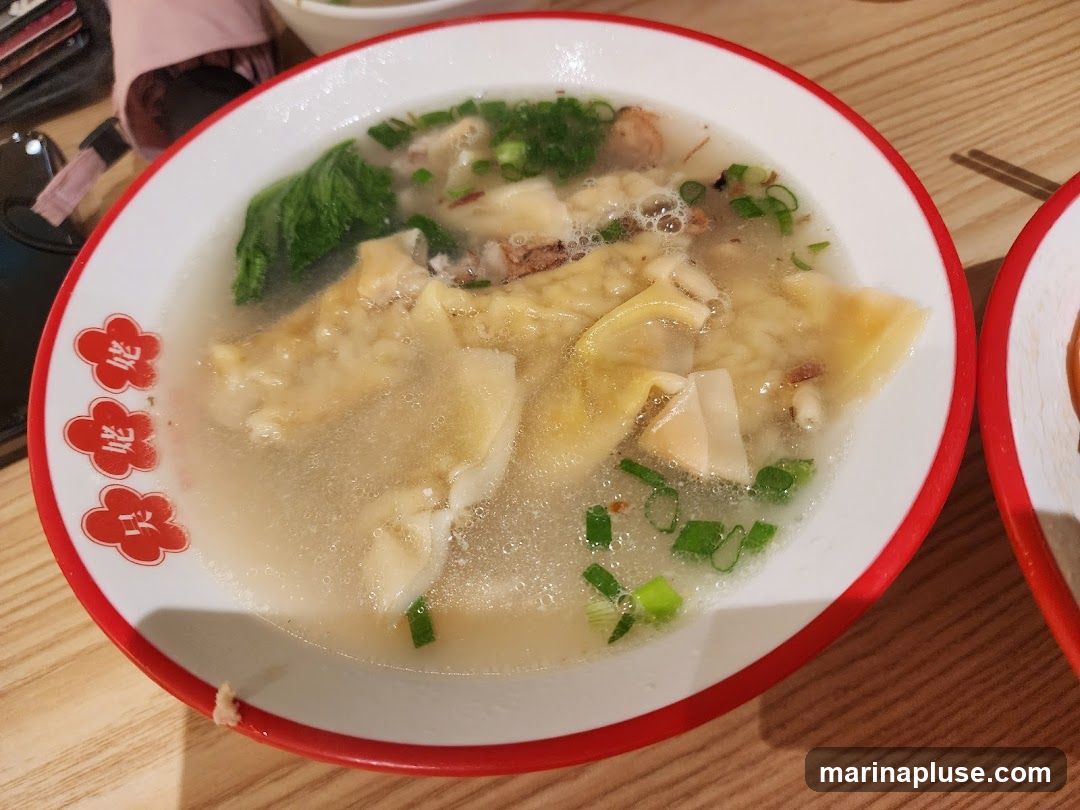 A close-up shot of a bowl of wonton soup. The soup contains wontons, green vegetables, and chopped scallions. The bowl has a red rim with floral designs. The table surface is made of light-colored wood.
