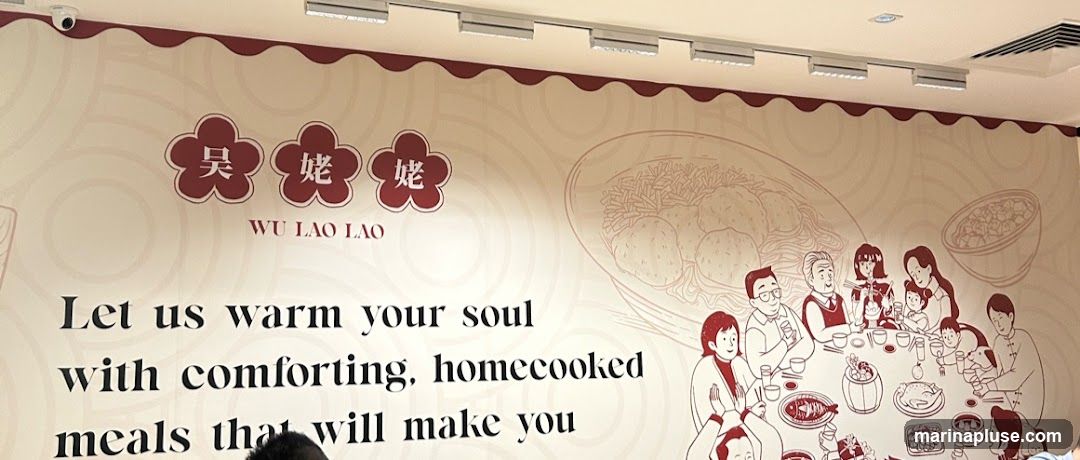 A mural on a wall features the text "WU LAO LAO" and the slogan "Let us warm your soul with comforting, homecooked meals that will make you". The mural also includes an illustration of a family gathered around a table laden with food, and drawings of dishes like a whole roasted chicken, fish, and dumplings.