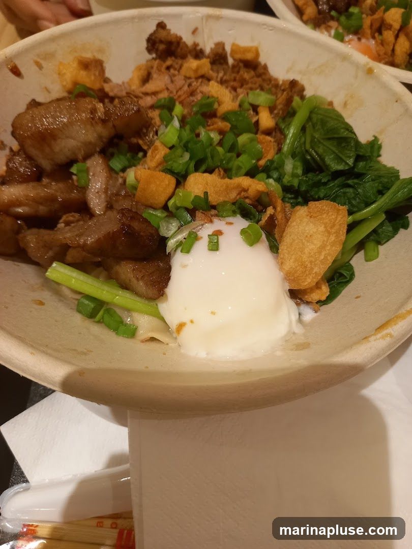 A close-up shot of a bowl of food. The bowl contains sliced pork, a soft-boiled egg, green vegetables, and crispy toppings. The food is served in a light-colored, disposable bowl.
