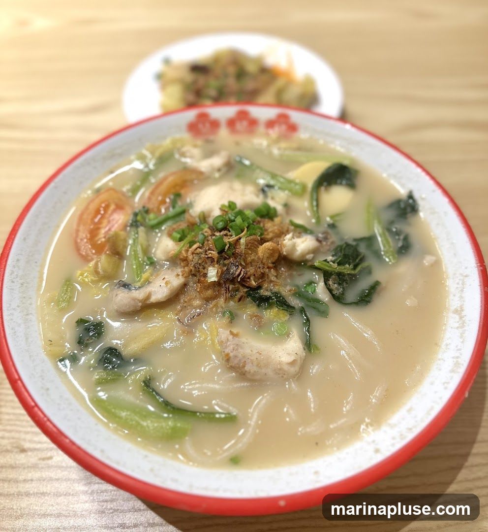 A close-up shot of a bowl of fish soup noodles, with pieces of fish, tomatoes, leafy greens, and tofu in a creamy broth. Fried shallots and chopped scallions are sprinkled on top. In the background, a plate of food is out of focus.