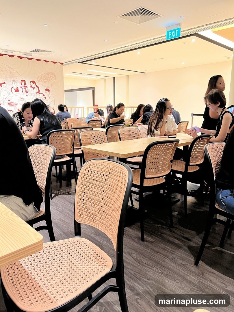 An interior view of a busy restaurant with several tables occupied by diners. The chairs have a woven cane design and are paired with light-colored wooden tables. A mural depicting people and food is visible on the wall in the background. An illuminated "EXIT" sign is mounted on the ceiling.