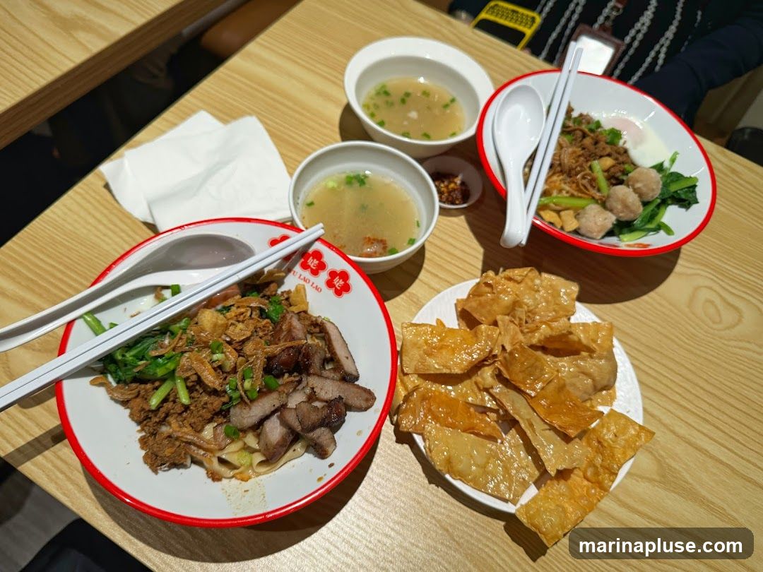 A close-up overhead shot of a meal on a wooden table. In the foreground, a white plate holds a pile of crispy, golden-brown fried wonton wrappers. To the left, a red and white bowl contains noodles topped with sliced roasted pork, minced meat, and green vegetables, accompanied by a spoon and chopsticks. Two smaller white bowls of soup and a small dish of chili flakes are also visible. In the background, another red and white bowl holds noodles with meatballs, greens, and a fried egg, also with a spoon and chopsticks.