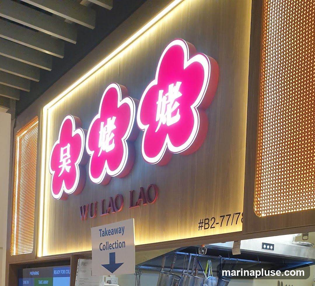 A close-up shot of a restaurant's illuminated signage. The sign features three large, stylized pink flower shapes with white borders, each containing a Chinese character in white. Below the flowers, the text "WU LAO LAO" is displayed in red. To the right of the sign, a code "#B2-77/78" is visible. A white sign with blue text indicates "Takeaway Collection" with a downward-pointing arrow. The background is a wooden panel with warm lighting.