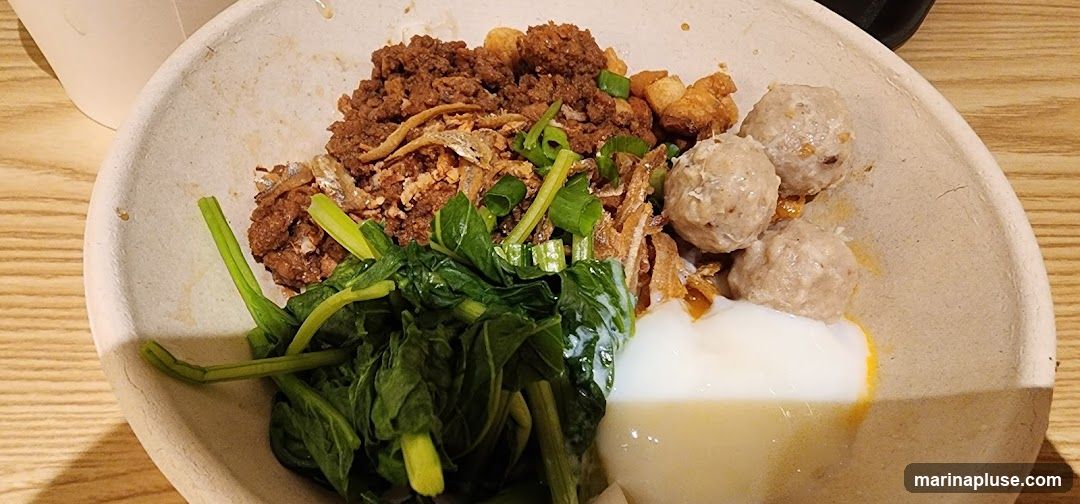 A close-up shot of a bowl containing noodles topped with minced meat, crispy fried shallots, small dried fish, chopped green onions, and blanched greens. Three meatballs are visible on the side, along with a dollop of white sauce, possibly egg or mayonnaise. The bowl is made of a light-colored, textured material, and it sits on a wooden surface.