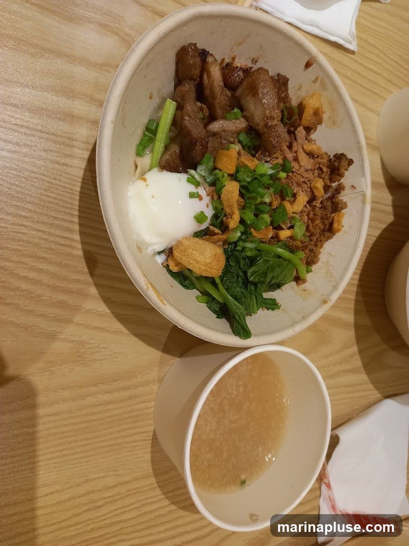 A bowl of noodles topped with sliced meat, a soft-boiled egg, chopped green onions, fried garlic bits, and greens. A small cup of broth is placed next to the bowl on a wooden table.