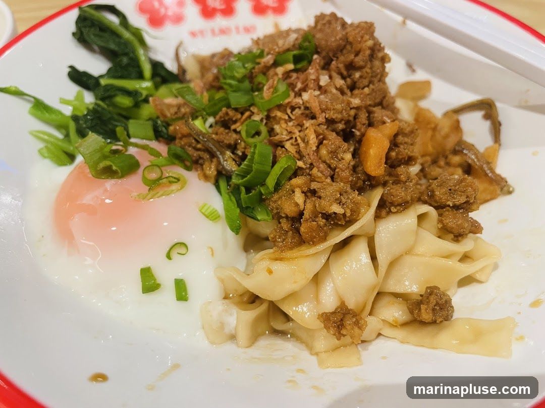 A close-up shot of a bowl of noodles topped with minced meat, a soft-boiled egg, and green onions. The noodles are wide and flat, and the minced meat appears to be seasoned. A side of leafy green vegetables is also visible. The dish is served in a white bowl with a red rim.