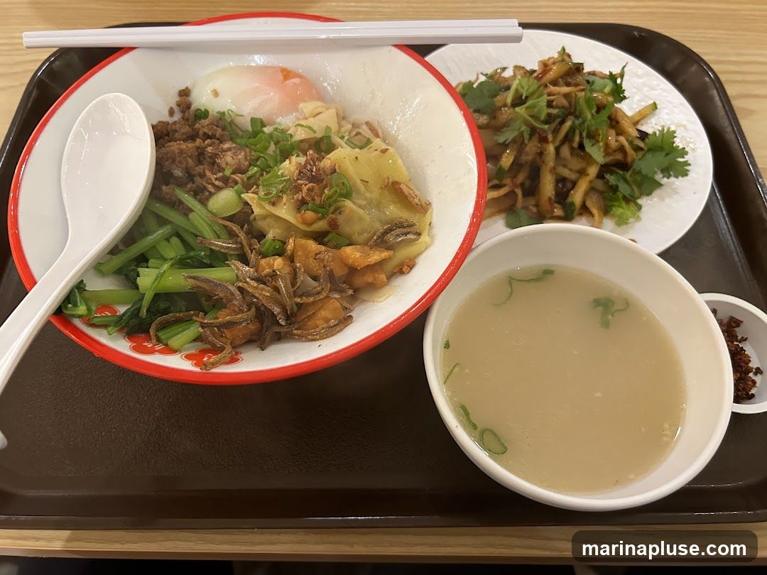 A bowl of noodles topped with a soft-boiled egg, minced meat, wontons, greens, and fried anchovies. A side dish of shredded vegetables with herbs and a bowl of broth are also present.