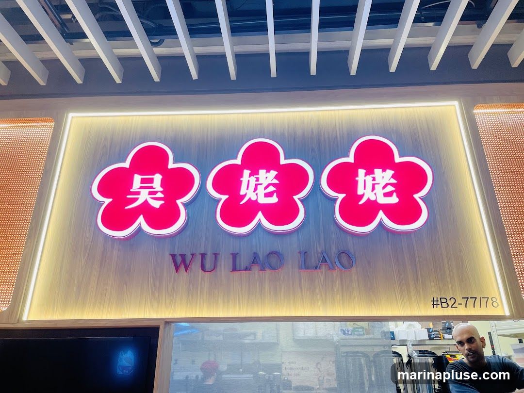 A close-up shot of the illuminated signage for "WU LAO LAO" with three red flower-shaped emblems above the text. The sign is mounted on a wooden-paneled wall. Below the sign, a man is visible behind a counter with some equipment.