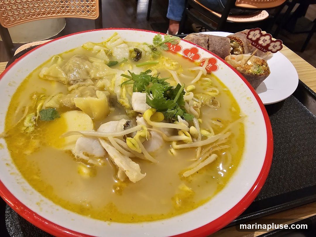 A close-up shot of a bowl of noodle soup with wontons, fish, bean sprouts, and greens. To the right, on a white plate, are two spring rolls, one cut in half. A small decorative flag with Chinese characters and "WU LIAO ZAO" is stuck into one of the spring rolls. The food is served on a black tray on a wooden table.